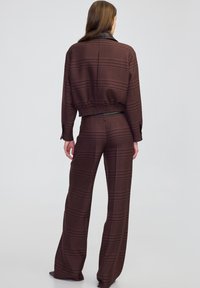 Una donna sta in piedi girata di spalle indossando una giacca a maniche lunghe coordinata con un motivo rosso scuro e nero e pantaloni larghi, su uno sfondo neutro.