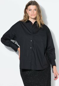 Camicia nera con bottoni, tasca, vestibilità ampia, maniche gather con lacci, tessuto morbido e superficie liscia. Presentata su uno sfondo bianco.