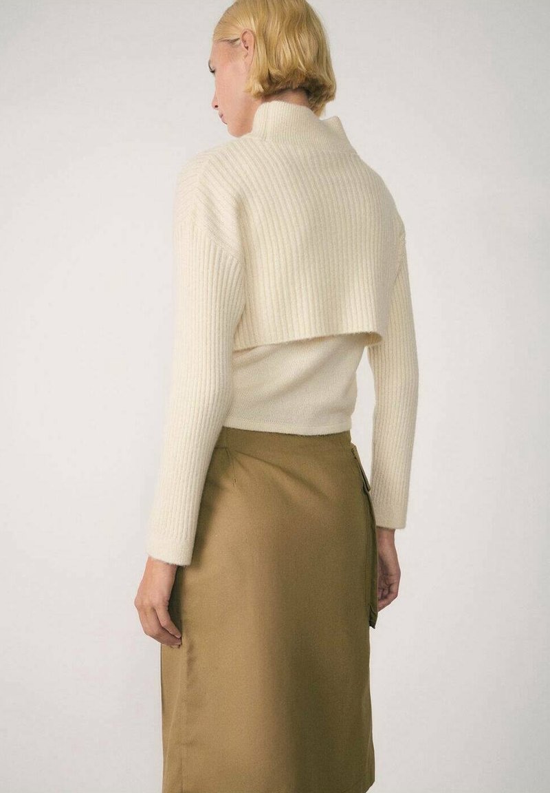 Pull en maille côtelée crème avec une coupe courte, associé à une jupe marron clair dotée d'une fermeture éclair latérale et d'une texture lisse. Vue de dos.