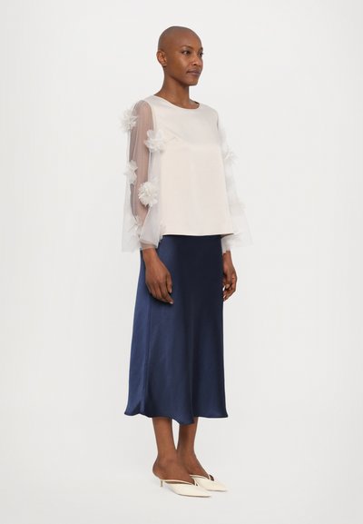Light-colored top with sheer, floral-embellished sleeves, paired with a flowing navy satin skirt and white pointed-toe footwear.