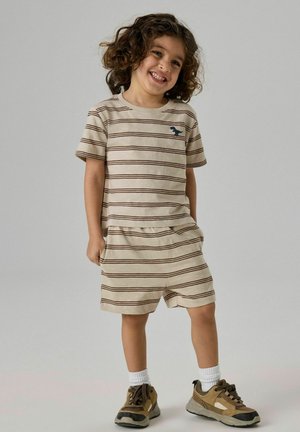 Naeratav laps lokkide juustega, kandes beeži triibulist T-särki ja sobivaid lühkseid pükse, seisab käed taskus, pruunid tossud ja valged sokid.