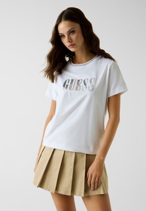 Jeune femme aux cheveux ondulés portant un t-shirt blanc Guess et une jupe plissée beige sur un fond clair uni.