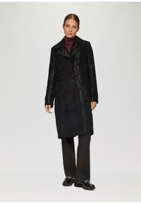 Manteau noir avec un motif texturé, large col et zip sur le côté. Porté sur un pantalon noir et des chaussures brillantes, offrant une silhouette longue.