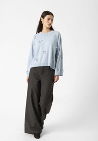 Model wearing a light blue sweater with decorative bows, paired with loose dark trousers, walking against a plain white background.