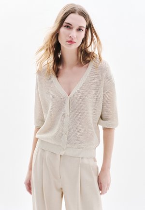 Mujer con cabello ondulado lleva un cárdigan suelto de punto de malla beige con botones y pantalones plisados crema de talle alto sobre fondo blanco.