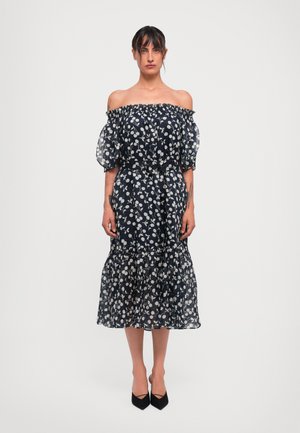 Gaun navy off-shoulder dengan motif bunga putih, lengan bervolume transparan, pinggang bagian yang pas, dan hem ruffle bertingkat. Sepatu hak tinggi hitam.