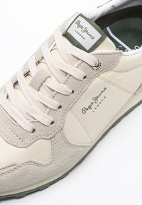 Béžové a světle šedé tenisky vyrobené z hladkého syntetického materiálu a semiše. Mají bílé tkaničky, zelenou logo nášivku a texturované detaily.