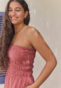 Robe sans bretelles, rose, avec smocks texturés et encolure à volants. Le tissu est léger, portée avec un collier en perles et des boucles d'oreilles créoles.