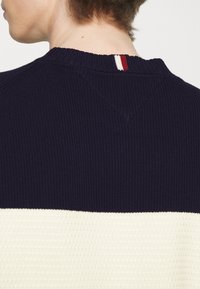 Maglione lavorato a maglia di un blu scuro con una striscia orizzontale bianca, caratterizzato da una superficie testurizzata e un accento rosso, bianco e blu al collo.