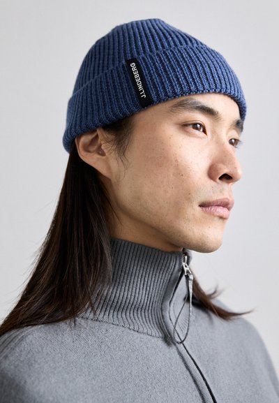 Bonnet en tricot bleu avec un côtes verticales et une étiquette noire, porté avec un pull gris à col montant avec une fermeture éclair.
