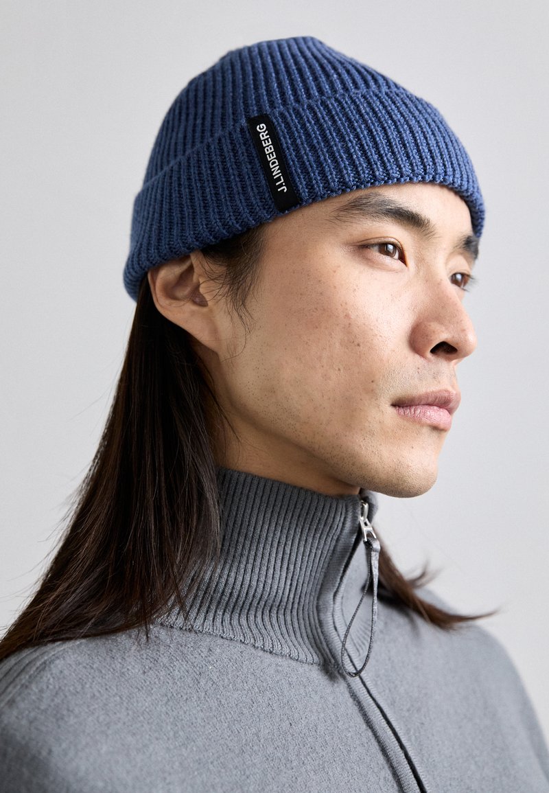 Gorro de malha azul com caneluras verticais e um rótulo preto, usado com um suéter cinza de gola alta com fecho de zip.