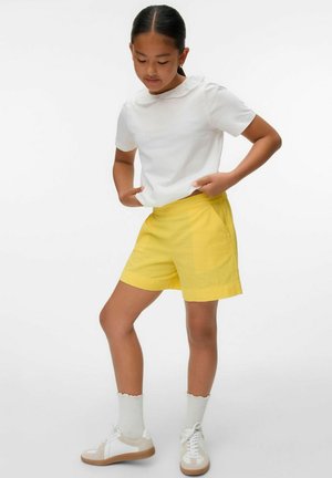 T-shirt en coton blanc avec un col en dentelle, associé à un short jaune. Le modèle porte des chaussures blanches avec des accents beiges et des chaussettes blanches à volants.