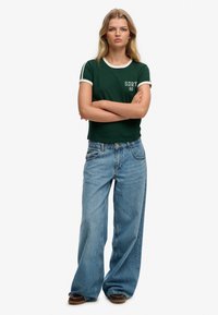 Groen T-shirt met korte mouwen, witte bies en opdruk "SDRY 82", gecombineerd met lichtblauwe, wijde denim jeans en bruine schoenen.