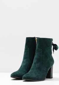 Stivaletti ankle in suede verde con punta a punta, zip laterale e nastro decorativo sul retro, dotati di tacco largo per stabilità.