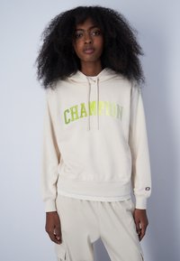 Felpa beige con la scritta "CHAMPION" in lettere verde metallico, cappuccio con cordoncino, polsini e vita a coste, realizzata in morbido tessuto.