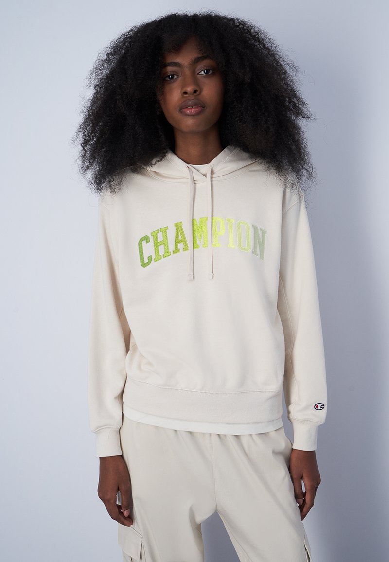 Felpa beige con la scritta "CHAMPION" in lettere verde metallico, cappuccio con cordoncino, polsini e vita a coste, realizzata in morbido tessuto.