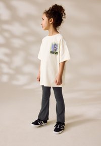 T-shirt blanc surdimensionné avec un design graphique comprenant des formes abstraites, associé à un pantalon slim gris foncé et des baskets noires avec des accents blancs.