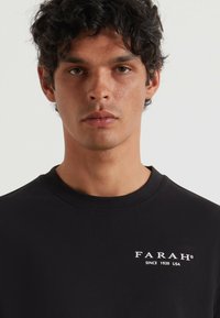 Czarny t-shirt z okrągłym dekoltem. Ma białe logo z napisem "FARAH" oraz "SINCE 1920 USA" po lewej stronie. Gładki materiał, dopasowany krój.