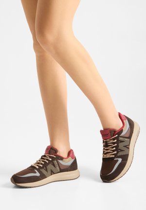Beine, die braune und rote Schnürschuhe mit beigen Sohlen tragen, stehen auf einem hellgrauen Boden vor weißem Hintergrund.