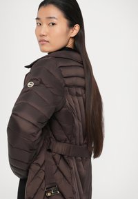 Veste matelassée marron avec une taille ceinturée, affichant un design matelassé et une patch de marque sur l'épaule gauche. Finition lisse, aspect léger.