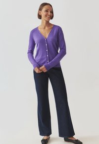 Purple cardigan with a deep V-neck, long sleeves, and button closure. Paired with high-waisted dark denim trousers and black shoes.