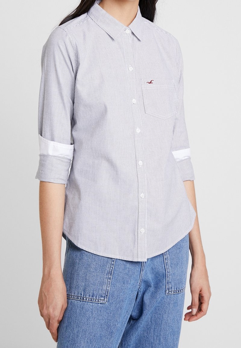 Chemise à boutons en tissu rayé bleu et blanc, avec une poche poitrine gauche, des détails de poignets blancs et une coupe décontractée.
