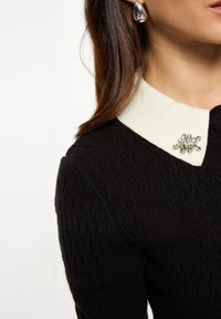 Suéter negro de punto trenzado con cuello blanco que presenta un adorno de broche de strass. Pendientes en forma de lágrima plateados completan el look.