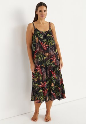 Kobieta o długich ciemnych włosach nosi czarną, bezrękawnikową sukienkę z nadrukiem tropikalnych liści w kolorach czerwonym, zielonym i beżowym, stojąc boso przy białej ścianie.