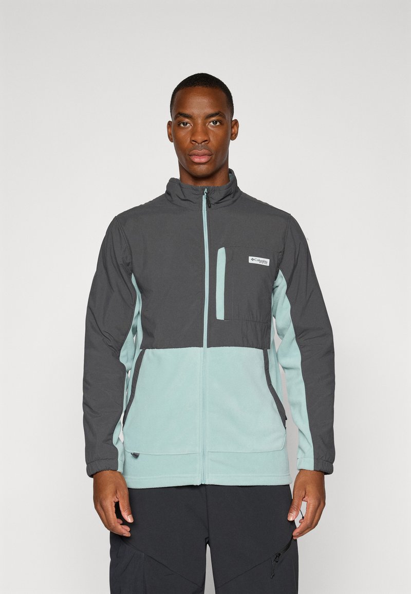 Chaqueta de hombre en gris oscuro y azul claro. Presenta un cierre de cremallera en la parte delantera, collar alto, bolsillo en el pecho y un diseño de paneles en contraste. Material suave, ajuste relajado.