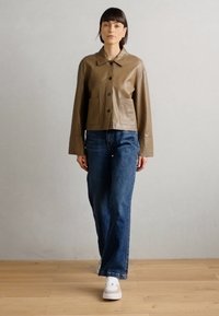 Veste en cuir marron raccourcie avec un col, fermeture à boutons sur le devant et deux poches avant, associée à un pantalon en denim bleu large et des baskets blanches.