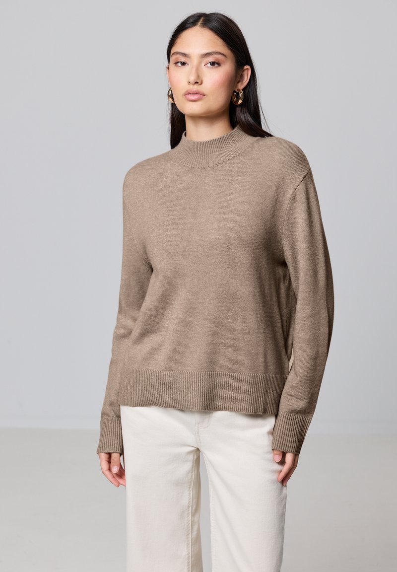Maglione lavorato a maglia color marrone chiaro con colletto a coste alto, spalle scese e orlo a coste, indossato sopra pantaloni a gamba larga color avorio.