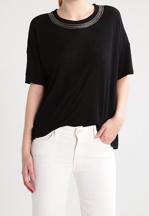 Mujer con una blusa negra de manga corta con bordados plateados alrededor del escote y unos vaqueros blancos de tiro alto, con una mano en el bolsillo.