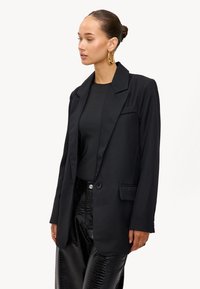 Zwarte op maat gemaakte blazer van een gladde stof, met een notch lapel, enkelvoudige knoopsluiting en zijzakken, gecombineerd met zwarte gestructureerde broek.