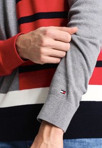 Pull en maille avec rayures rouges, grises et bleu marine. Les poignets sont côtelés ; écusson logo sur la manche. Le matériau semble doux avec une texture lisse.