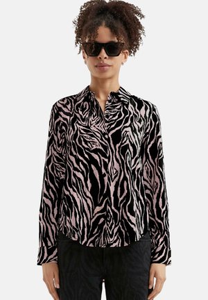 Femme aux cheveux bouclés portant des lunettes de soleil noires et un blouse à rayures zébrées noir et rose à manches longues avec un pantalon noir.