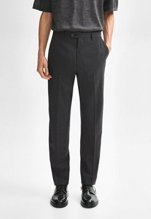 Homme portant un pantalon gris foncé ajusté avec un pli marqué à l'avant et des chaussures de ville noires vernies, debout devant un fond blanc uni.