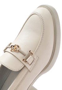 Mocasín de cuero beige con herrajes dorados decorativos y acentos elásticos. Punta redondeada y textura suave, con costuras blancas en contraste.