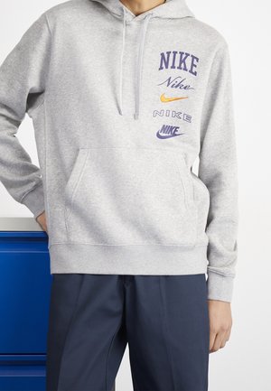 Personne portant un sweat à capuche Nike gris clair avec plusieurs logos Nike et un pantalon bleu marine, debout avec une main dans la poche près d'une surface bleue.