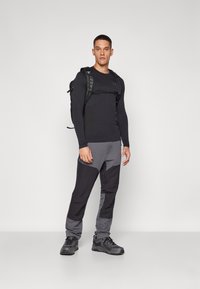 Schwarzes Langarmoberteil, grau-schwarze farbblockierte Hose und schwarze Wanderschuhe. Inklusive einem schwarzen Rucksack mit Tragegurten.