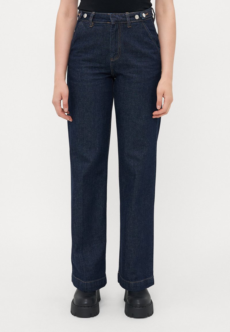 Woman wearing dark blue straight-leg jeans and black chunky boots, standing against a plain light background.
