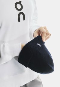 Person wearing white sweatshirt holding a navy blue knitted beanie hat with a small fabric logo tag.