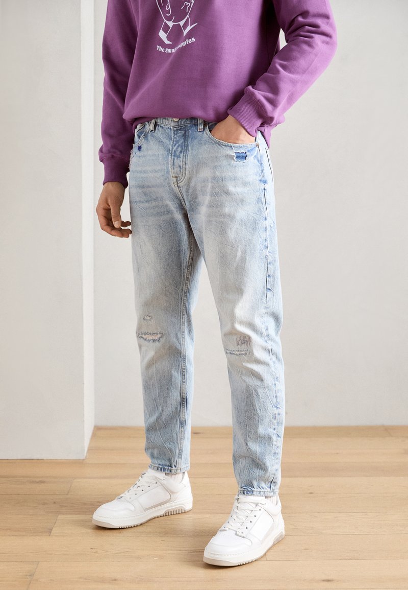 Lys blå denimjeans med en avslappet passform, med slitasje og rifter. Kombinert med hvite joggesko og en lilla hettegenser.