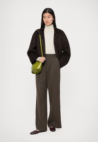 Jachetă maro cu fermoar, pulover crem cu gât, pantaloni maro cu talie înaltă, pantofi maronii, geantă verde tip crossbody rotundă. Texturi netede și croială ajustată.