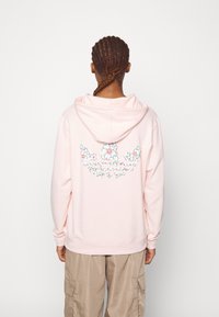 Sudadera con capucha rosa hecha de tela suave con un bolsillo tipo canguro. Bordado floral en la parte trasera con detalles en verde y rojo. Ajuste relajado.