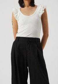 Vit ribbad tanktopp med volangärmar i genomskinligt tyg, kombinerad med svarta vida byxor. Påtagliga detaljer inkluderar mjuk textur och åtsittande design.