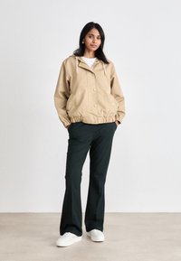 Beige, hettejakke med elastiske mansjetter og frontknapper, paret med mørkegrønne bukser og hvite joggesko. Glatt tekstur, avslappet passform.
