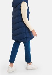 Gilet imbottito blu navy con cappuccio, caratterizzato da cuciture a zig-zag. Abbinato a un maglione chiaro e jeans blu, completato da sneakers bianche.