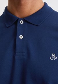 Tmavě modré polo tričko z texturované tkaniny, s klasickým límečkem, třemi bílými knoflíky a malým bílým vyšívaným logem.
