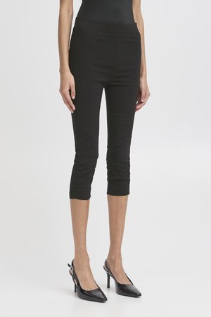 Nő fekete, szélesített szárú, ráncolt szegélyű nadrágot és fekete hegyes orrú, pántos magas sarkú cipőt visel, egyszerű háttér előtt áll.