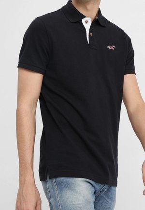 Černé polo tričko s bílým límečkem, se malým červeným logem. Vyrobeno z bavlny, s krátkým rukávem a mírně přiléhavým střihem.